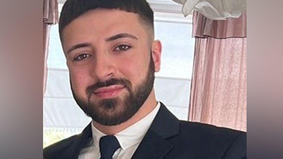 Head and shoulders photo of Kyle Clifford smiling at the camera wearing a black jacket and tie and white collared shirt.