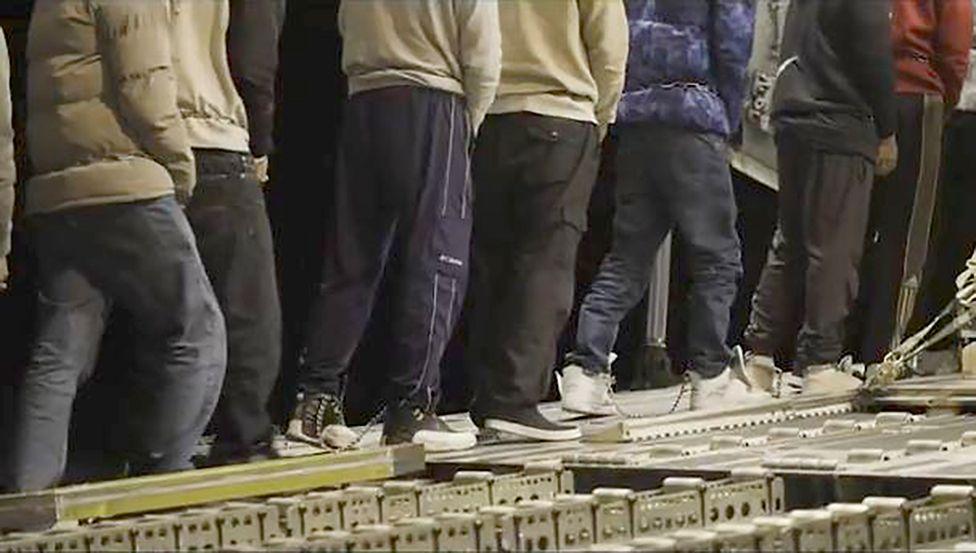 A still from a video produced by US Border Force showing migrants in casual clothes and warm coats with their legs chained together, walking up a ramp onto a military aircraft. The image is cropped to conceal their faces and to highlight the chains around their ankles.