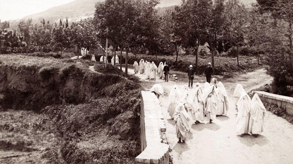 Tlemcen, Algeria. A black and white 19th Century photo showing white-clad women walking back from a cemetery with some crossing a bridge and others walking along a tree-lined road. Behind the first set of women are three men in dark clothing with (possible) Fez hats