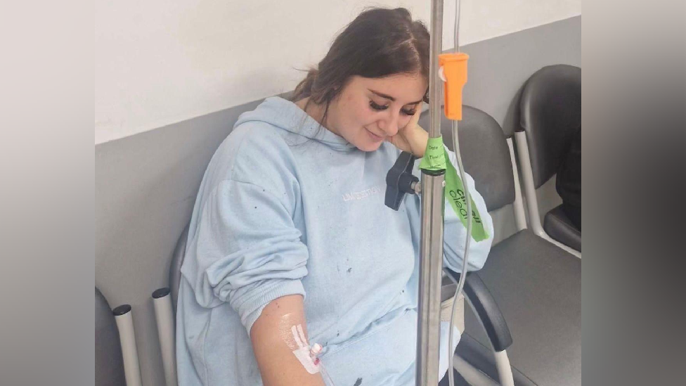 Paige Roberts sitting down in a hospital chair having IV fluids administered. She is wearing a light blue hospital gown and is looking down at the floor