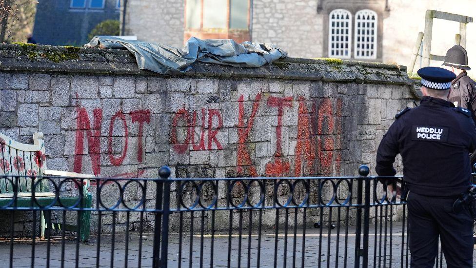Grafiti i lyer me sprej të kuq që thotë "Jo Mbreti Im" në murin jashtë Katedrales së Shën Asafit. Dy oficerë policie mund të shihen duke parë grafitin.