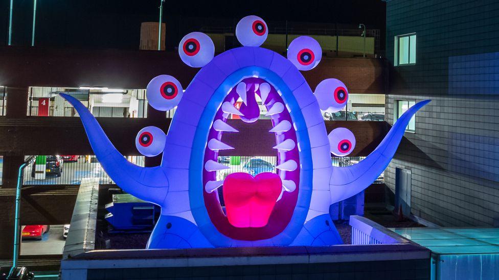 A blue inflatable monster with a gaping mouth, spiky teeth and seven eyes stands on a rooftop in front of a car park