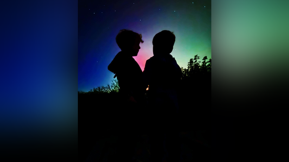 The silhouettes of two young children right in the center of a blue, green and pink Northern lights lit sky