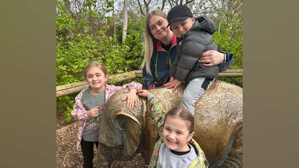 The family are all smiling at the camera for a posed photograph. Louis is sitting on a statue elephant and his mother Leigh has her arms around him. Evie and Paisley are standing next to the statue.