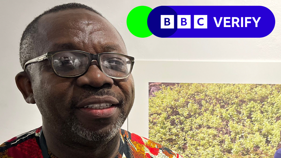 A head and shoulders portrait of Dr Leo Igwe as he looks at the camera wearing a colourful shirt, with the corner of a photo hung on a wall behind him just visible, showing shrubbery. 