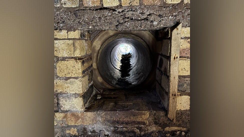 A circular ventilation tunnel leading from inside a bunker to the outside world. It is set inside an aged brick wall that has cracks in it