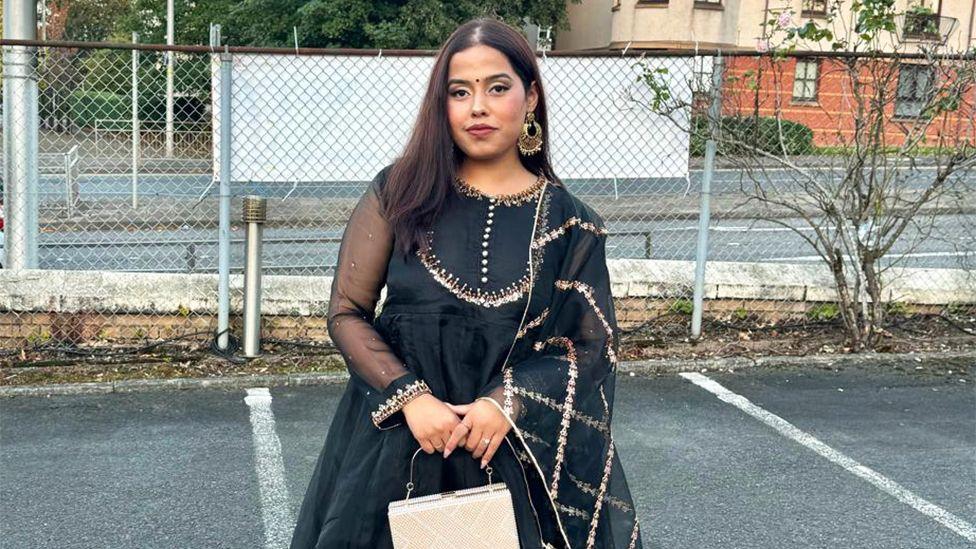 Taniya, a female, wearing a shiny patterned black coloured traditional clothing with a cream coloured purse, standing in a car park bay, with a silver fence behind her.