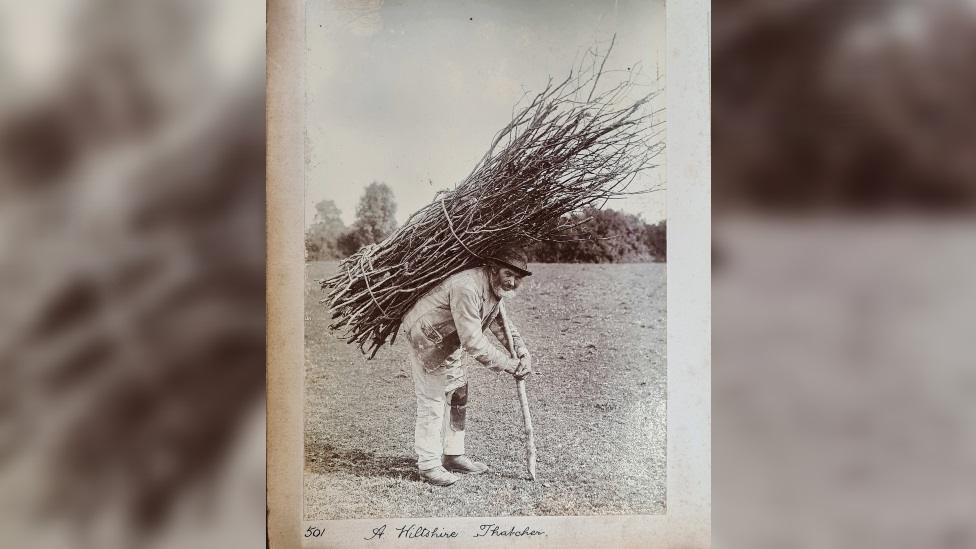 Black and white photo of a thatcher. He has a grey beard and weathered face and is stooping. His hands hold a pole supporting a bundle of hazel on his back.