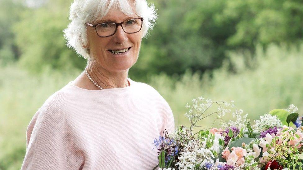 A bespectacled Gill Hodgson wearing a pink top and holding a large bunch of flowers standing in an outdoor location with trees in the background.