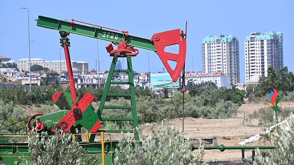 A red and green oil pump at an oil well in the capital city Baku, with apartment blocks, a screen and a park in the background 
