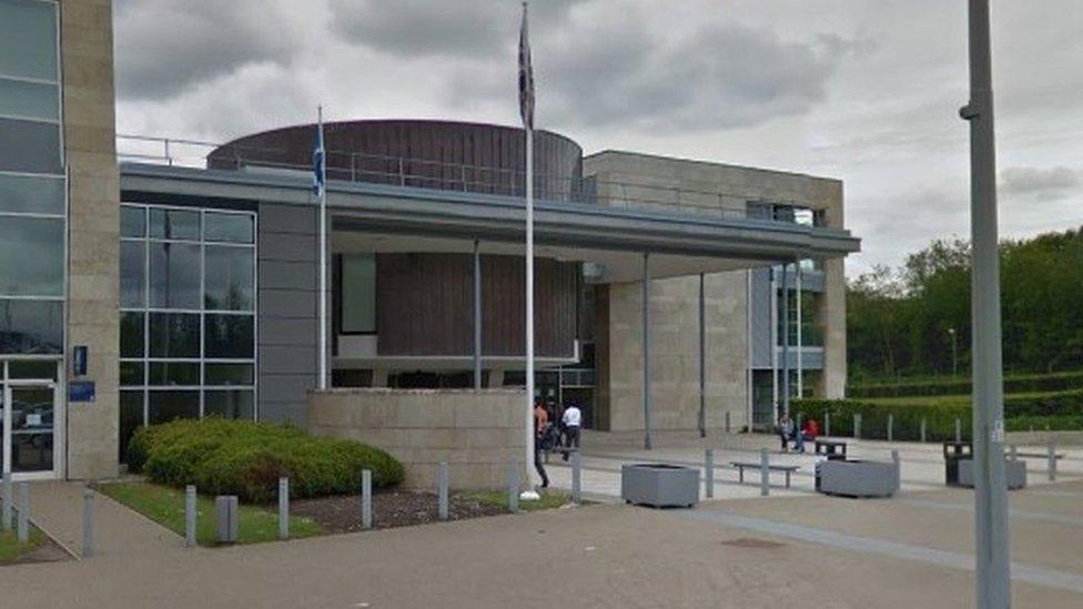 Grey and brown court building in a pedestrianised area. There are people, flagpoles, seats, planters and bushes outside