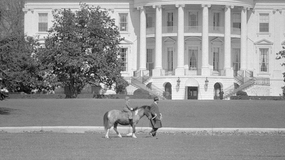 With a Secret Service man leading the way, Caroline Kennedy, the President's daughter, takes a ride on her pony, Macaroni, in 1962