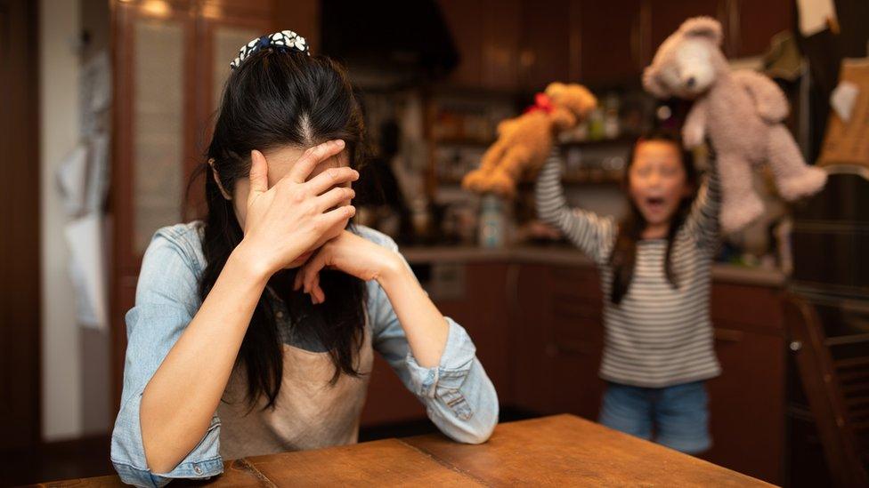 A woman with her head in her hands and a lively child behind her