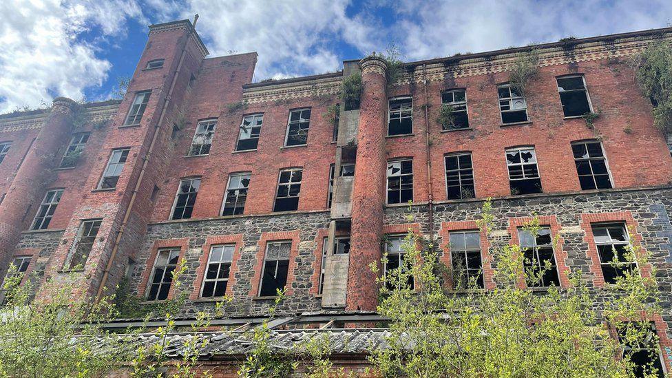 A side view of a derelict multi-storey building at Hilden Mill in the aftermath of an arson attack in May 2021.  Several windows of the red brick building are broken and there are plants growing from the windows and gutters. 