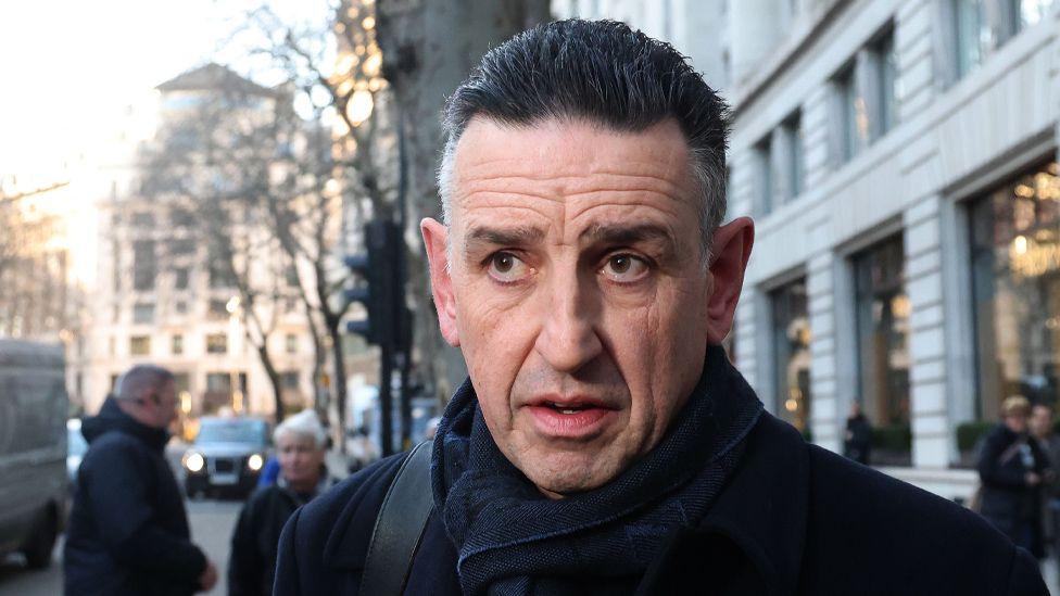 A headshot of William Paul Patterson outside the Post Office Horizon IT Inquiry. He is wearing a black overcoat and behind him is a street with people in the background.