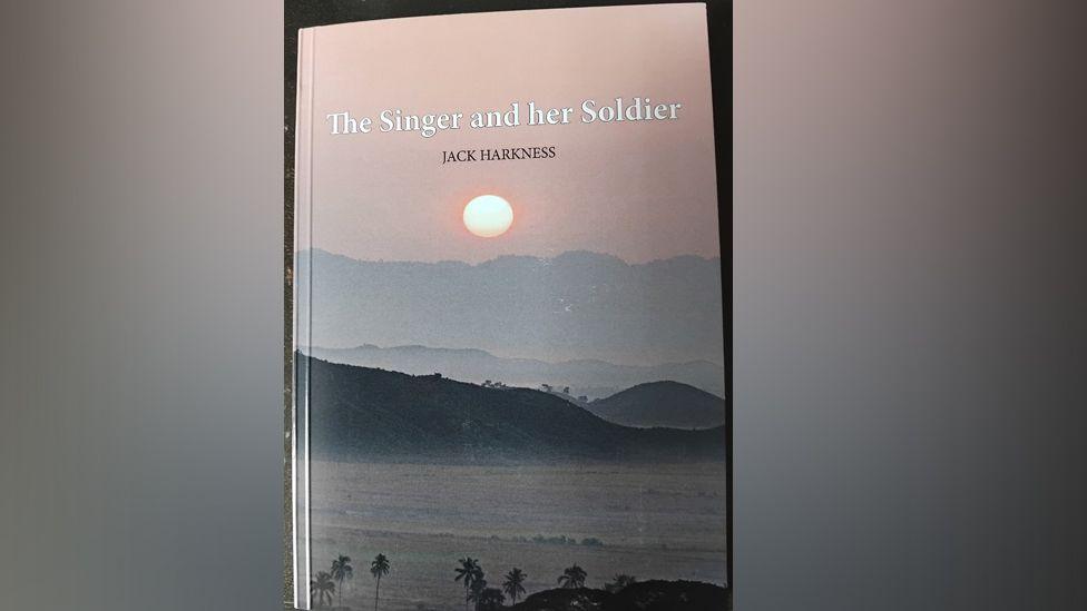 A photo of the front cover of the book. It says The Singer and Her Soldier in white font with the name JACK HARKNESS in black capitals below that. The photo shows sunset over a tropical sea, with palm trees in the foreground and hills and mountains behind the waves. A pale pink sky with a glowing sun is at the top.
