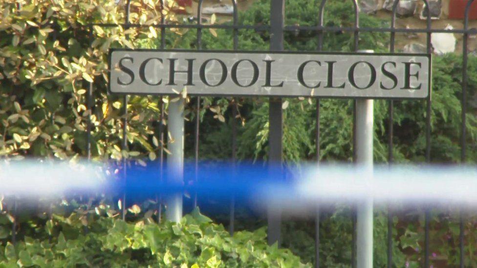 Road sign stating "School Close" in High Wycombe where the attacks happened. In front is a police blue and white cordon and behind greenery and a metal fence.