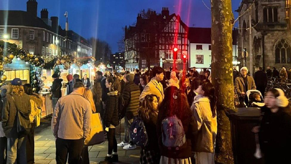 Tens of people stand around York Christmas Market at night time.