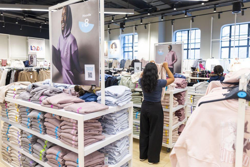 A woman stands in a Primark store, holding up a hoodie. You can only see her back. She is surrounded by rows of neatly folded clothes