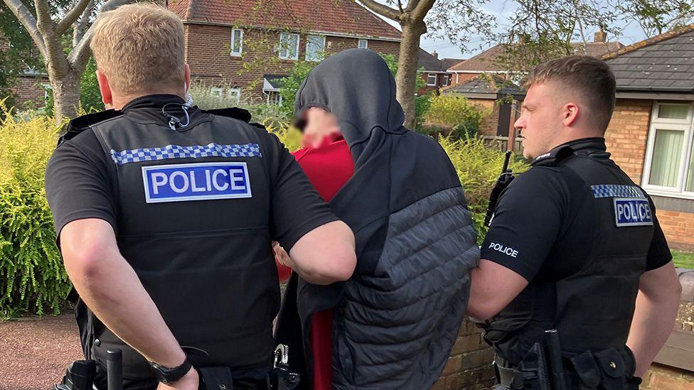 A close up of police escorting a man down a street. The man's face is blurred and he has a tracksuit top over his head.