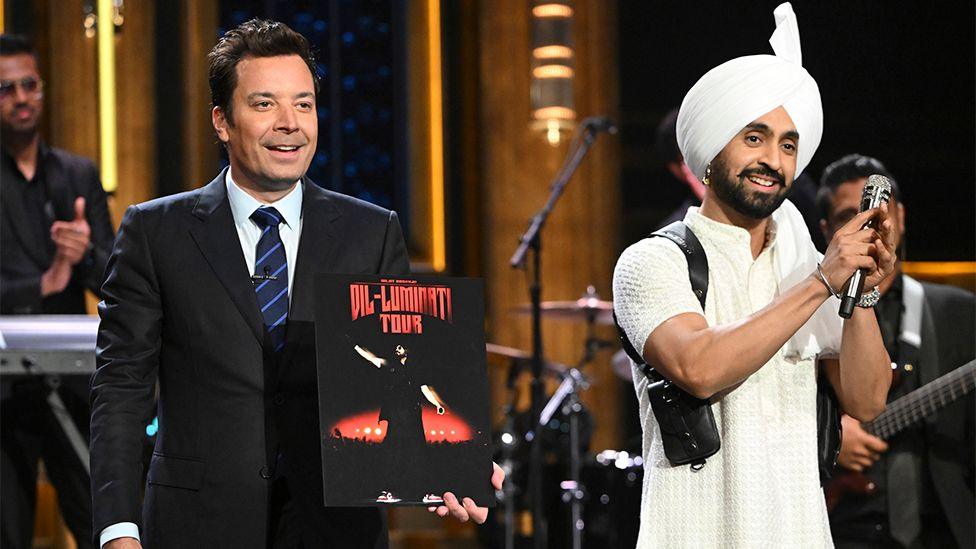 US host Jimmy Fallon wearing a black suit and white shirt with Diljit Dosanjh, who is wearing white with a black microphone in his hand, as he makes a pray gesture with his hands towards the audience.