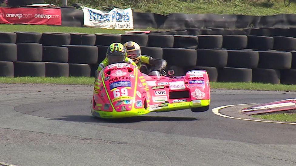 Nigel Limb in the side car of a pink motorcycle covered in sponsorship logos sits behind the rider as the bike comes round a corner with the sidecar wheel off the ground in front of a bank of stacked tyres. Both are wearing yellow racing leathers and helmets