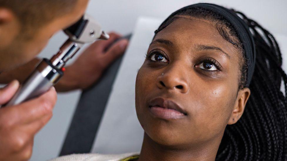 A stock image of a woman being treated at the opticians. The optician is looking into an implement that is scanning the woman's eye. The woman has long, black braided hair.