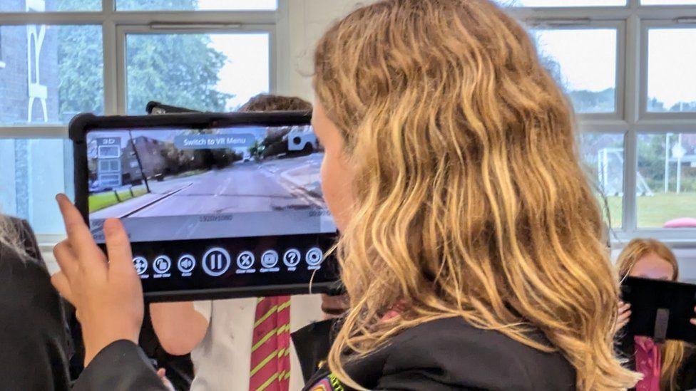 A student in a blue blazer holding one of the tablets watching a VR bike ride showing a road approaching a junction