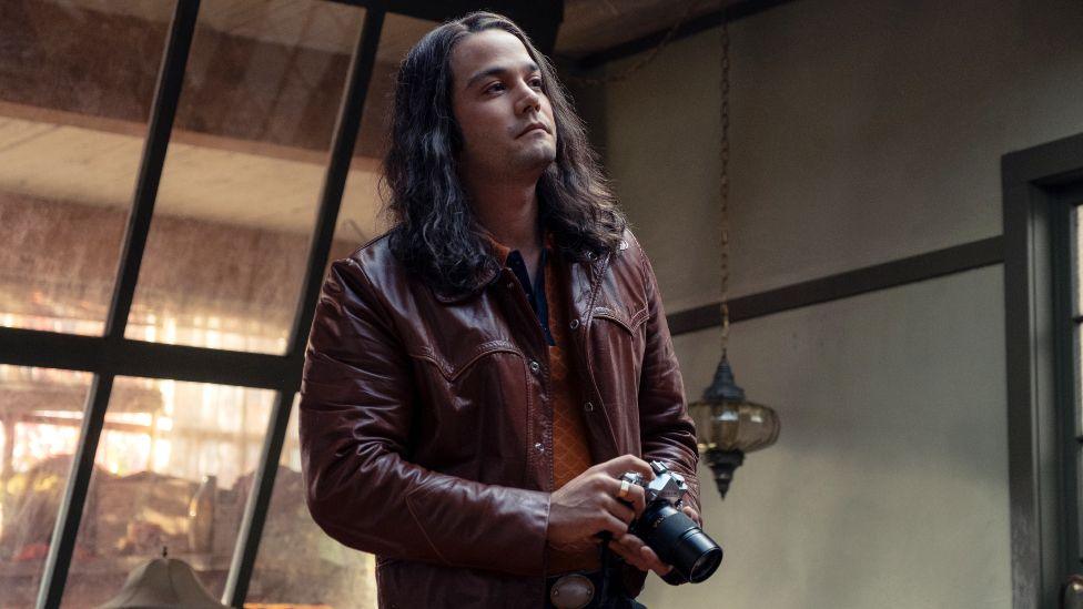 Daniel Zovatto as Rodney Alcala in Woman of the Hour. He has long hair, a burgundy leather jacket and is holding a camera.