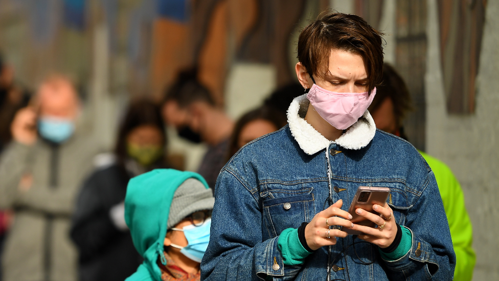 A young man wearing a mask checks his phone while queueing for a Covid test in Melbourne