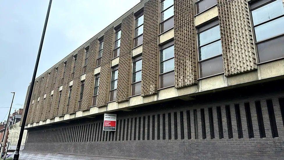 A three-storey building with patterned concrete slabs along its front - each placed either side of a window. There is a red and white marketing sign fixed to the side of the building.