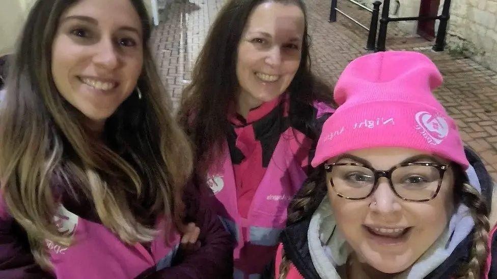 Three women pictured outside at night. They are stood close together, all smiling at the camera. They are all wearing bright pink high-vis vests, and one is wearing a pink beanie with Night Angels embroidered on it. 