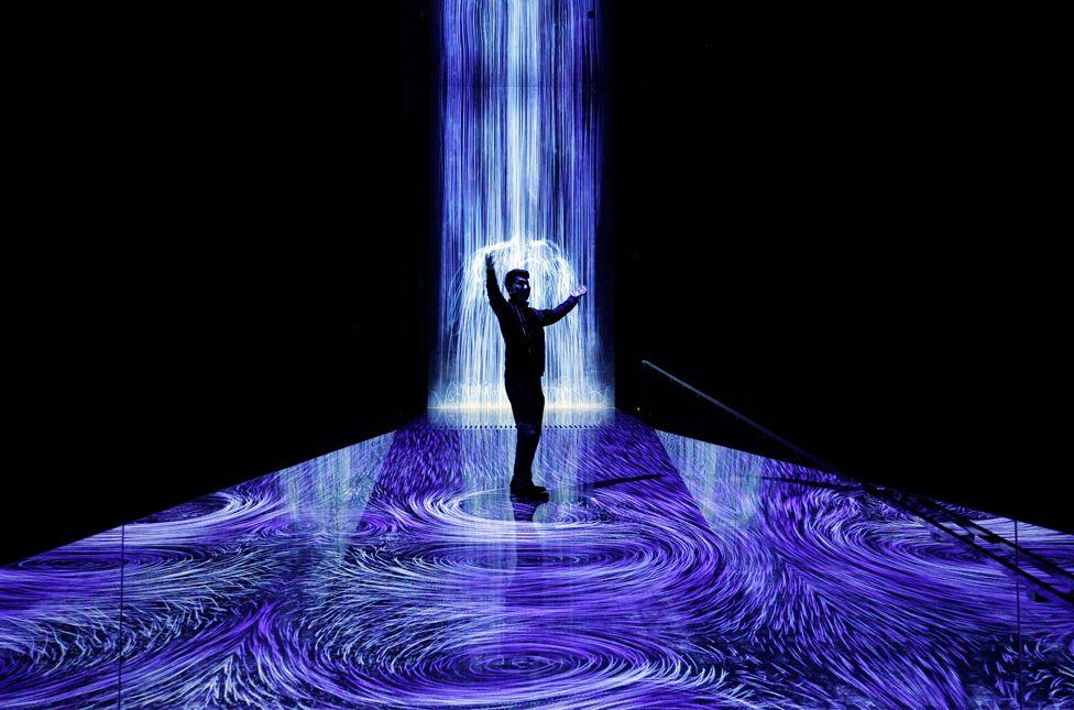 A media member poses at the 'Existence in the Flow Creates Vortices' area during a press preview at team Lab Planets Tokyo in Tokyo, Japan, 20 January 2025. A person, in silhouette, poses at a sound and light installation. Blue light to resemble rain fall and puddles is projected in a black room.
