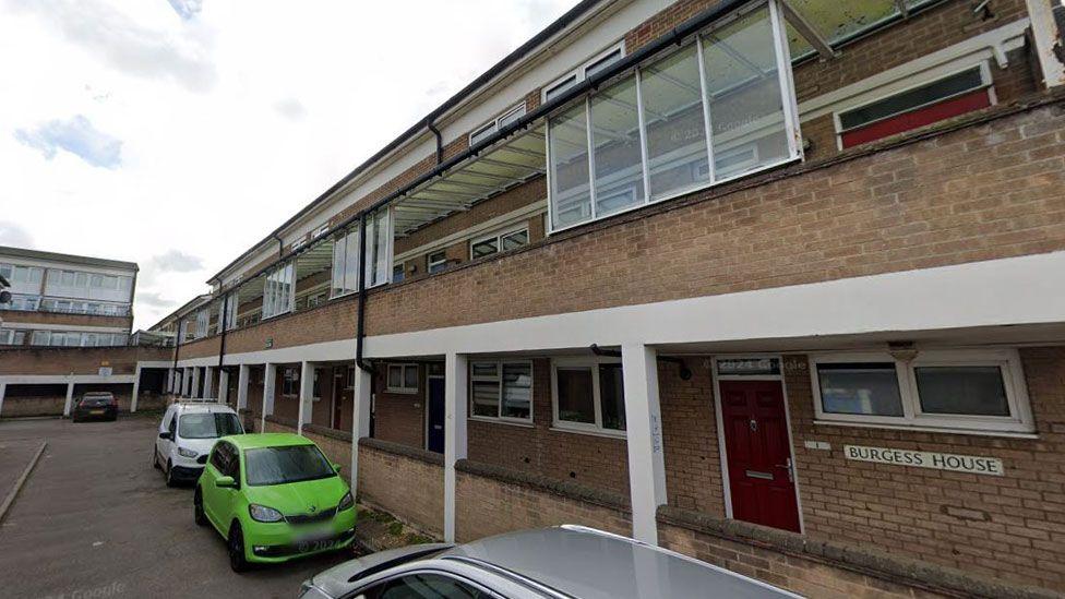 A 1970s block of flats in Cambridge. It is a three storey block made of brick. The ground floor has a walkway covered by the balcony along the first floor. Cars are parked in front of the block. 