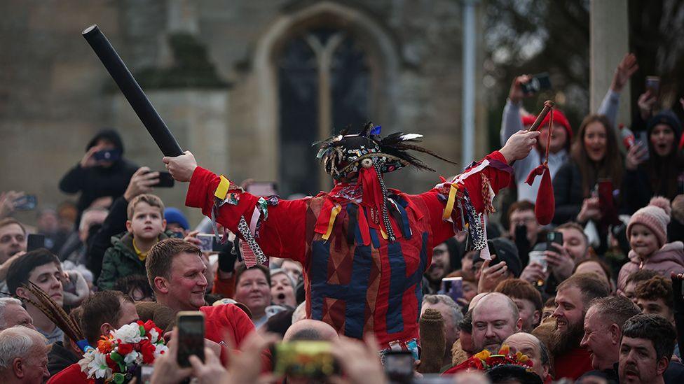 The fool delivers his opening speech surrounded by villagers. He wears a red jacket with strips of fabric and an a hat adorned by feathers. He holds a black baton in one hand and a purple velvet bag on a string in the other.