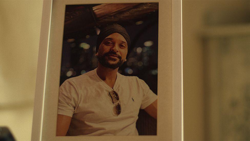 A picture of Bobby, from the Netflix show, in a white rectangular frame where is wearing a white tshirt with sunglasses tucked into his top.