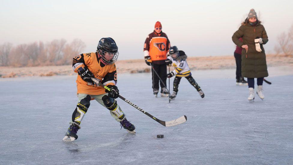 A man and two children under 10 playing ice hockey on a frozen field at Upware, Cambridgeshire. One child is in front, wearing an orange top, shin pads, a black helmet with a face guard and is reaching towards a puck. A second child is behind, also in action, and behind them both is the man in orange ice hockey gear and to their right is a woman in a brown coat on white skates