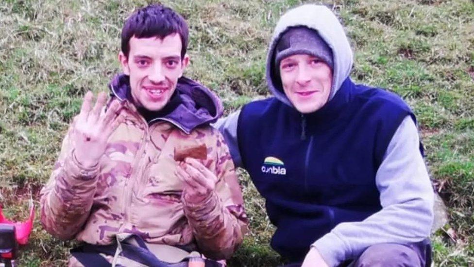 Josh holds up an axe head and four fingers sitting next to his brother Stephen