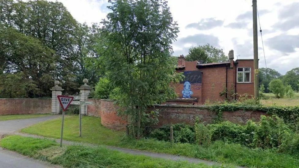 A red brick building behind a wall with large shrubs and graffiti showing