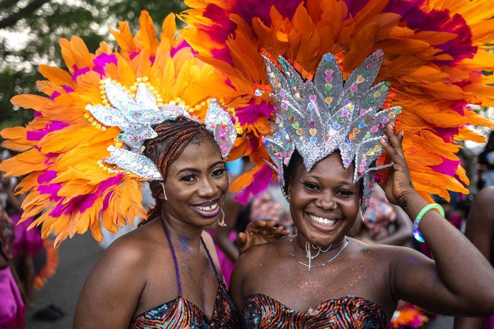 Two women in headdresses made of silver material and orange and pink feathers smile at the camera at the carnival in Calabar, Nigeria