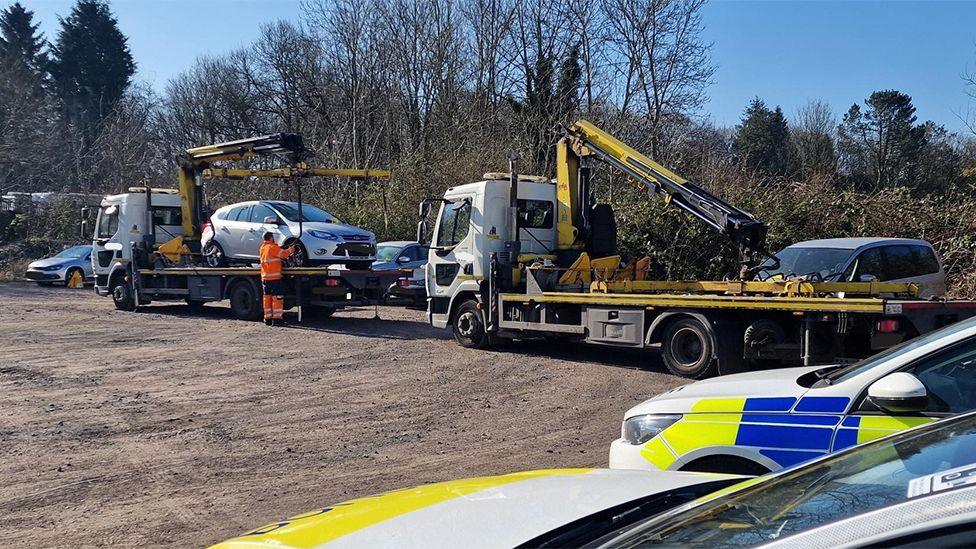 Cars being put on to recovery trucks so they can be towed away. In the background is a clamped car and in the foreground are two liveried police cars