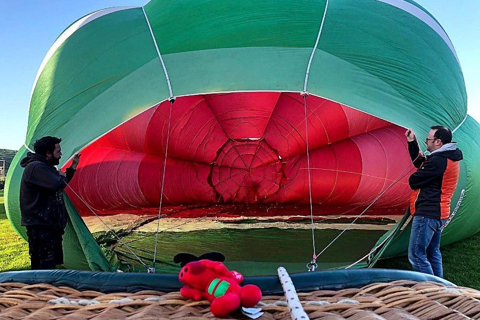 A wicker balloon basket is on its side in the foreground of the photo and the picture is taken from the top of that side. The viewer is looking into the interior of the balloon envelope lying partially inflated on the ground. It is green with one band of white visible at the top, and the interior and top is red. Two men wearing jeans and winter jackets stand either side of the envelope holdding it up. Various cords attach the basket to the balloon. There is a red Welsh dragon toy attached to the side of the basket in the centre of the picture at the bottom.