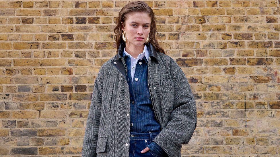 Female Topshop model wearing a dark grey winter coat over a dark blue pinstriped shirt and jeans, standing in front of a brick wall