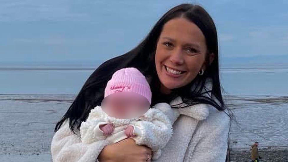 Kiena Dawes with long black hair smiling wearing a cream furry coat holding her baby dressed in a pink woolly hat, white furry coat and pink leggings and pink boots on the promenade with the sea in the background