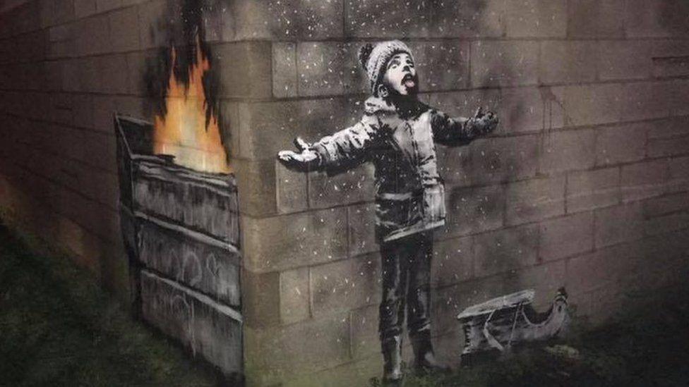 Banksy drawing across two walls at right angles - the right side shows a young child with his arms splayed, a sled at his feet and tongue out looking at what looks like falling snow. The left side reveals the snow is in fact ash from a fire burning in a dump truck.