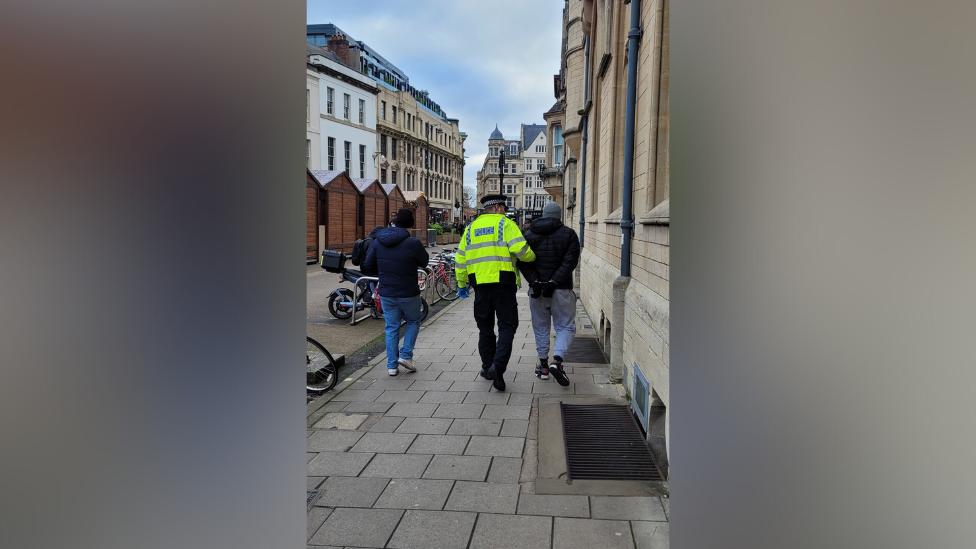 A man is led away wearing handcuffs by a uniformed officer