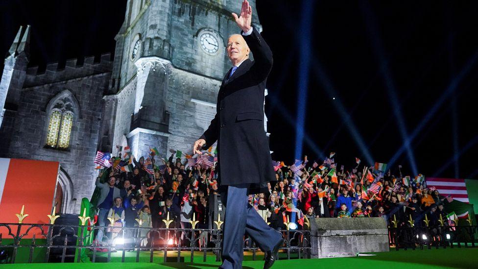 US President Joe Biden, wearing a blue suit and a dark blue overcoat, walks on stage on a green carpet and in front of a crowd of people waving Irish and US flags during an event at the grey-stone built St. Muredach's Cathedral in Ballina