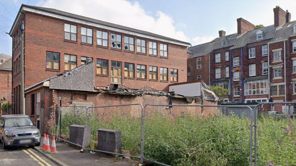 Lanson House, on Mount Street in Preston, is a large three-storey rectangular redbrick building with many broken windows next to a fenced-off area with overgrown weeds.