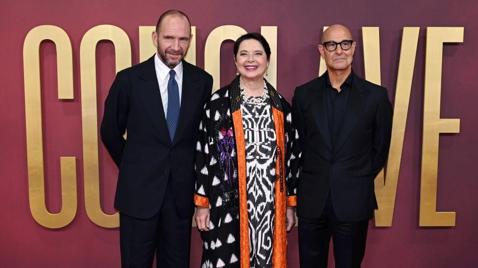 Ralph Fiennes, Isabella Rossellini and Stanley Tucci stand in front of a purple wall with large gold text that says "Conclave"