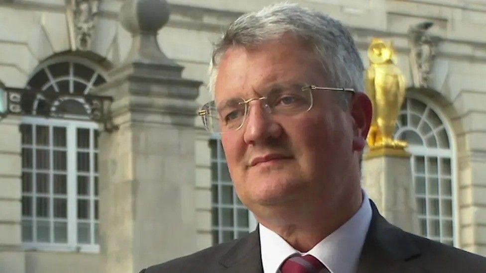 Leeds Council leader James Lewis - a man with grey hair and glasses. He is wearing a suit and stood against the backdrop of Leeds City Hall.